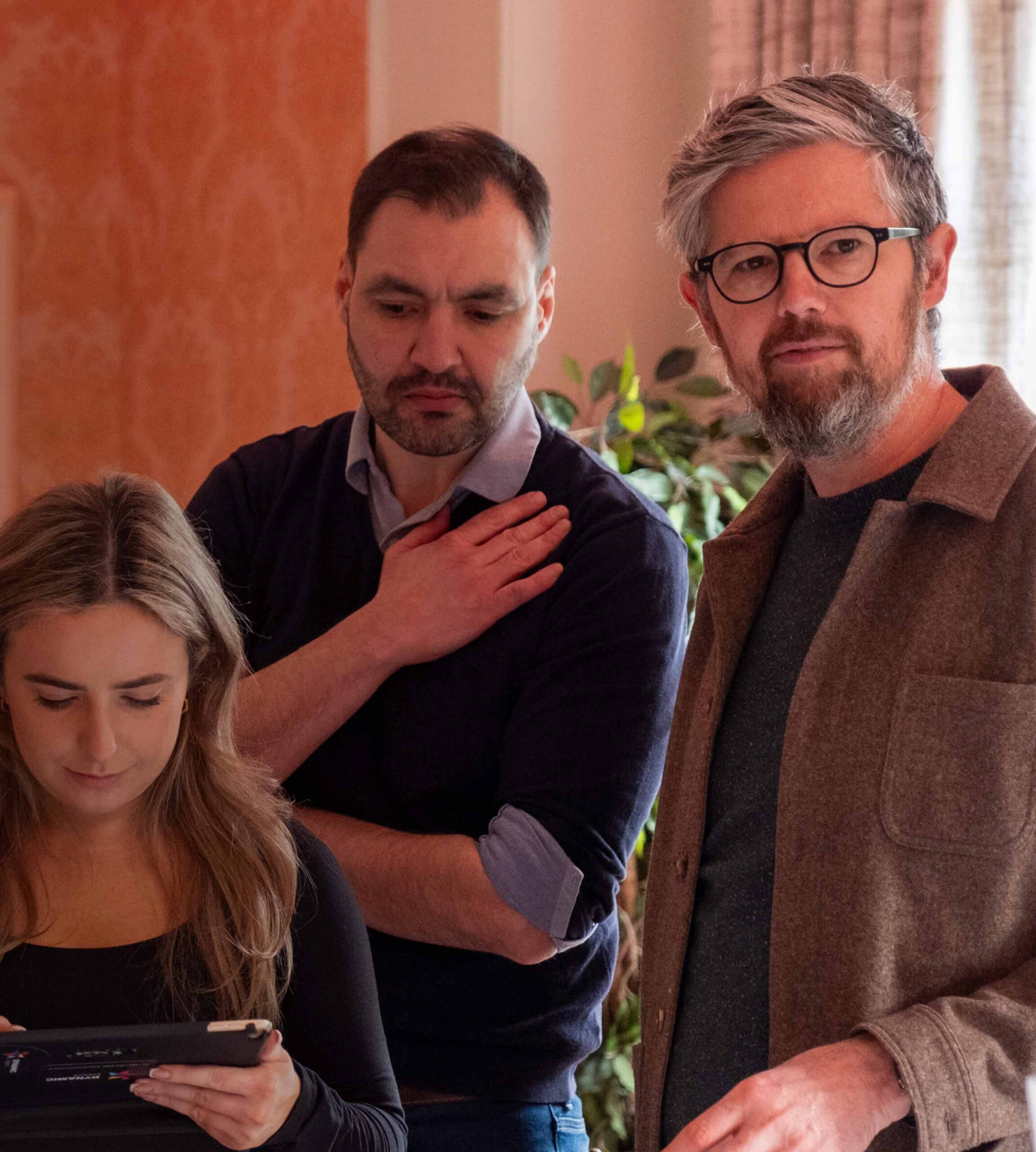 Three people indoors: a woman holding a tablet and looking down, while two men stand nearby, one with his hand on his chest. They appear to be engaged in discussion or observation. Warm lighting and a plant are in the background.