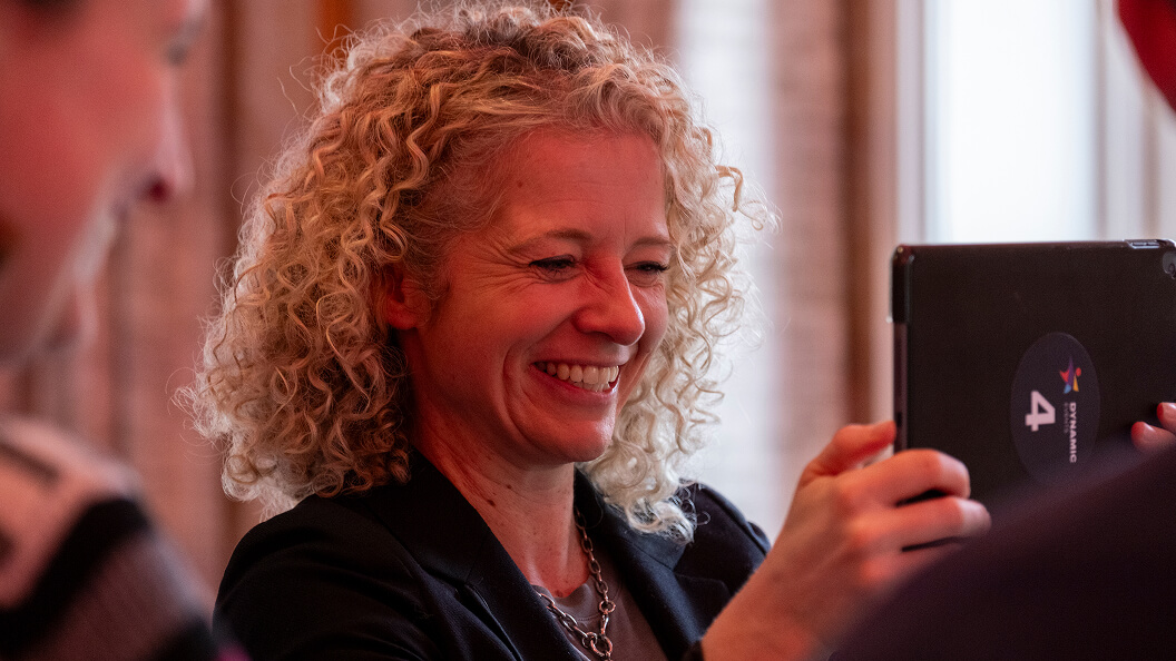 A smiling woman with curly blonde hair holds up a tablet to take a photo, with another person partially visible in the foreground. The background is softly blurred.