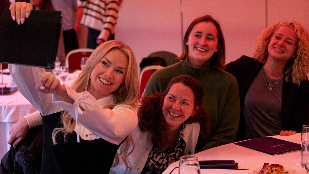 Four women sitting at a table smile and pose for a selfie together at an indoor event, with papers and drinks on the table and other people in the background.