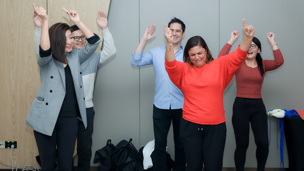 Five people stand indoors, smiling and raising their arms as if dancing or celebrating. They appear to be enjoying an informal or team-building event. The background is a plain wall with some bags and cables visible on the floor.