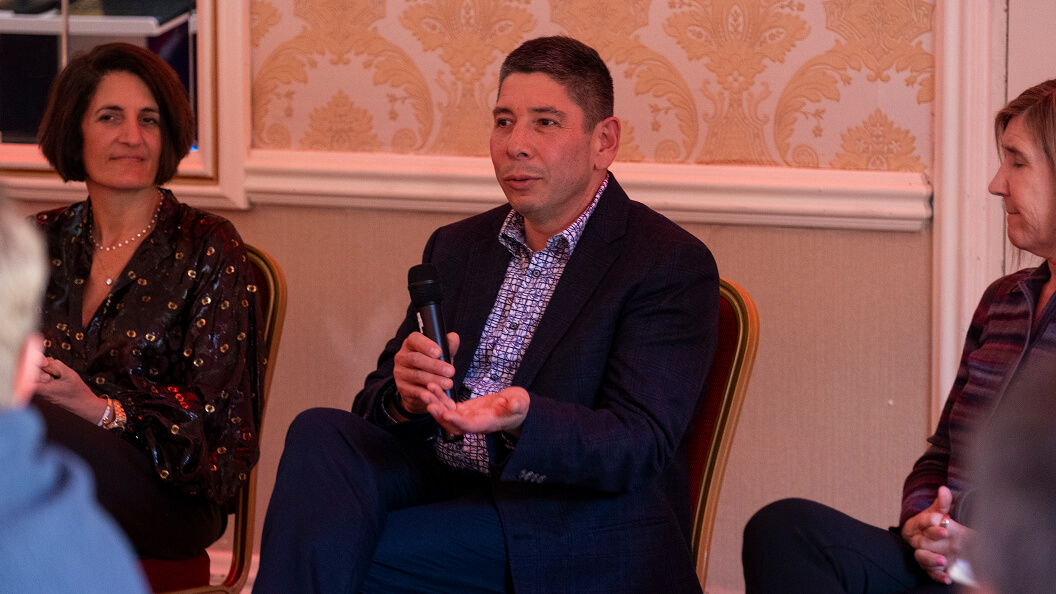 A man in a suit speaks into a microphone while seated between two women during a panel discussion in a room with ornate wallpaper.
