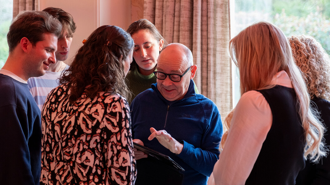A group of six people gather closely indoors, looking at a digital tablet held by an older man in glasses and a blue sweater. They appear engaged and focused on the screen. Natural light comes from a window behind them.