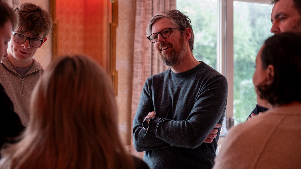 A man with glasses and a beard, wearing a dark sweater and watch, stands with arms crossed, smiling while talking to a small group of people indoors near a window.